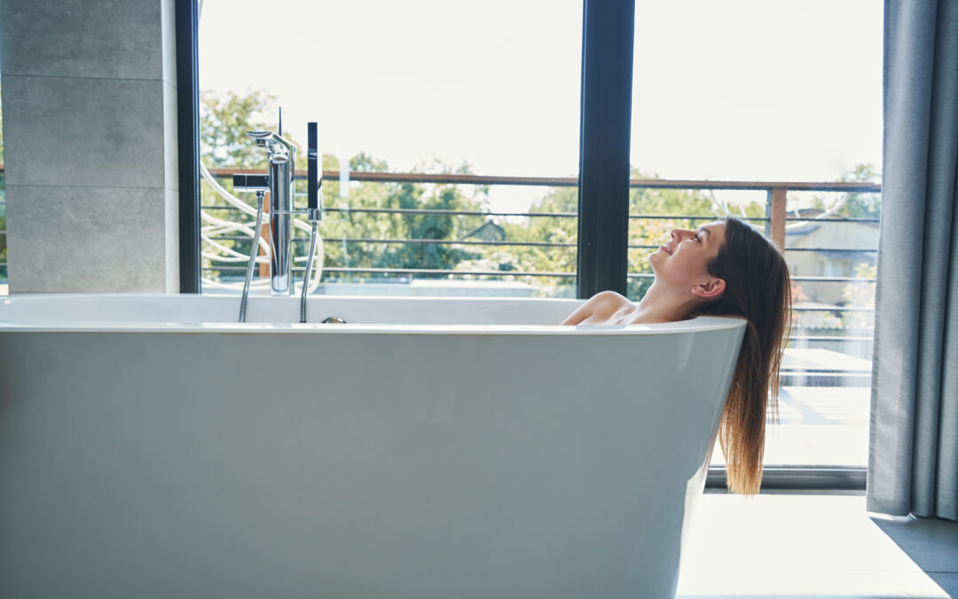 Woman relaxing in a modern freestanding bathtub with morning light streaming through large windows.